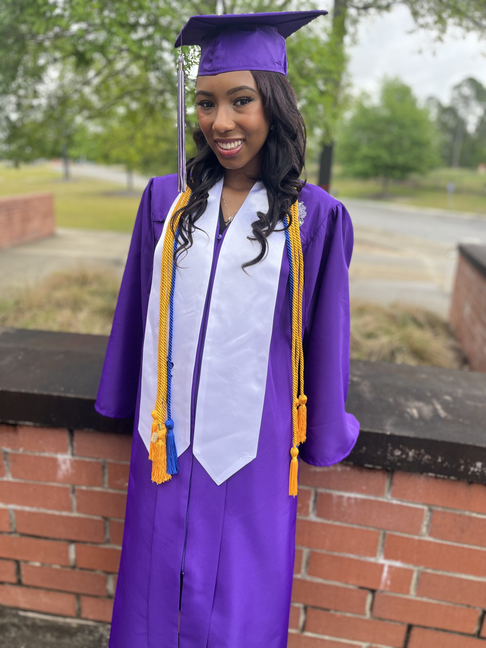 Me in purple cap and gown leaning on red brick